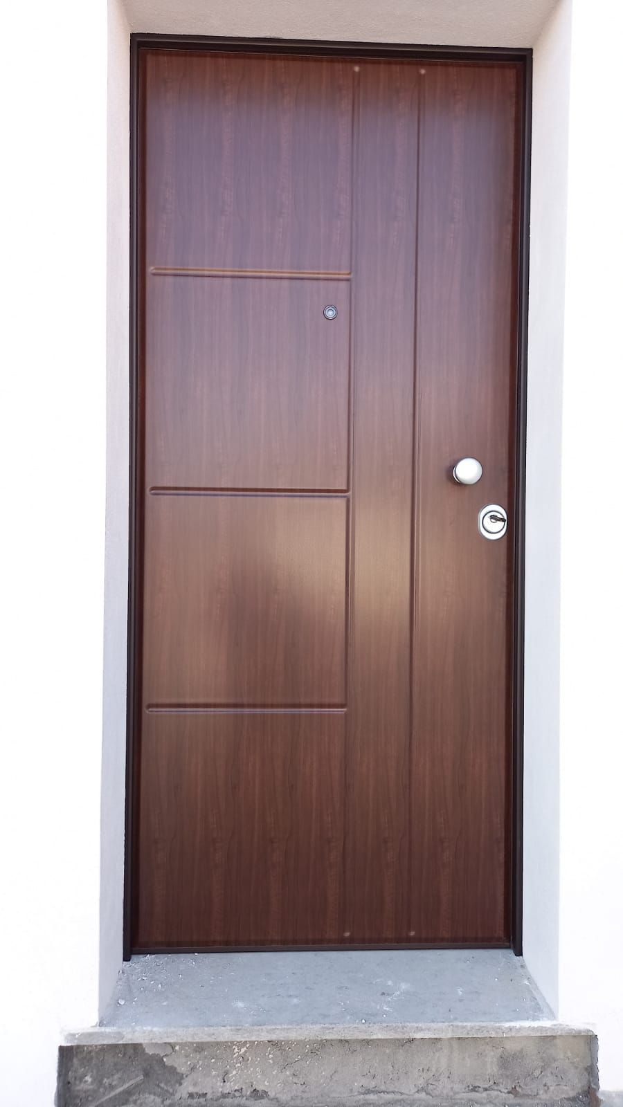 Porta d'ingresso chiusa in legno marrone con maniglia e serratura argentate, incastonata in un muro bianco sopra un piccolo gradino di cemento.
