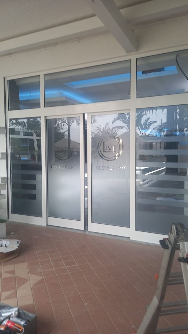 Ingresso del negozio con vetrina in vetro, pannelli laterali satinati e portico piastrellato sotto una tettoia.