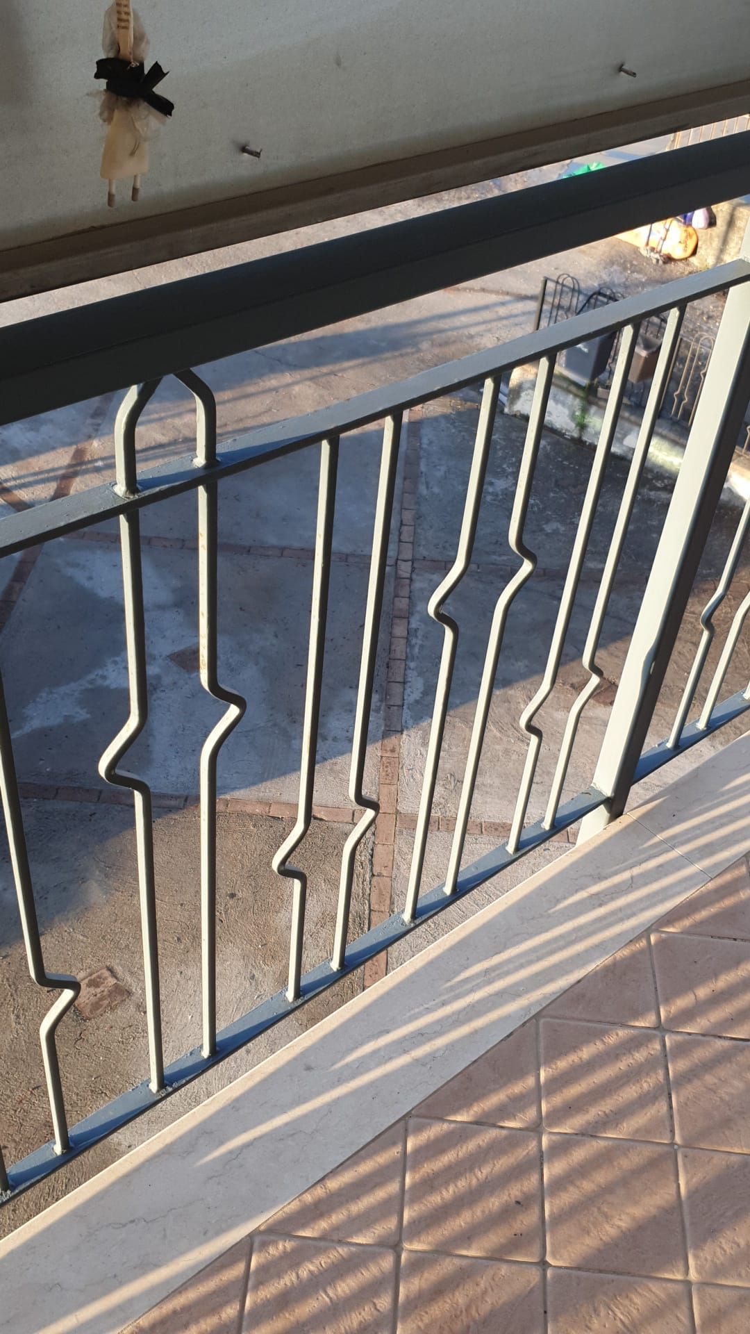 Ringhiera del balcone in metallo nero esposta alla luce del sole, con pavimento piastrellato e vista sulla strada sullo sfondo.