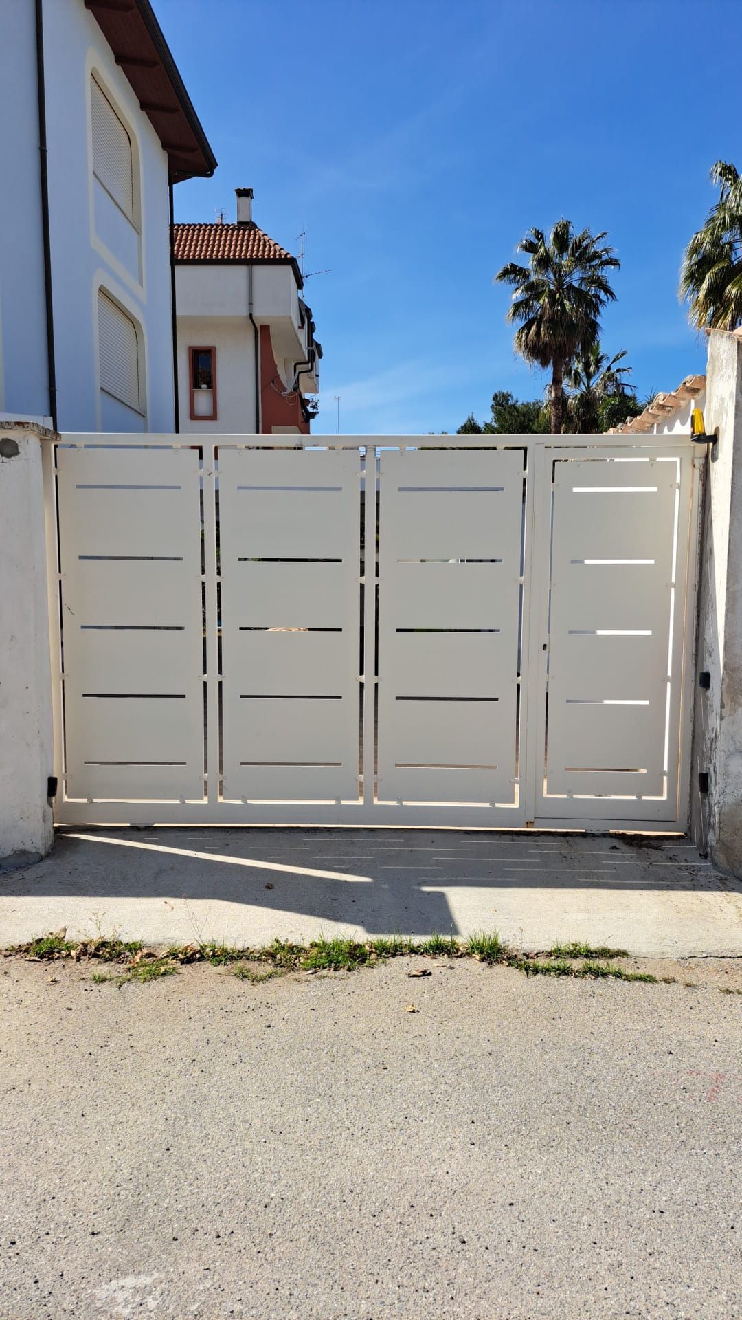 Cancello carrabile in metallo bianco tra le case di una strada soleggiata, con una palma sullo sfondo.