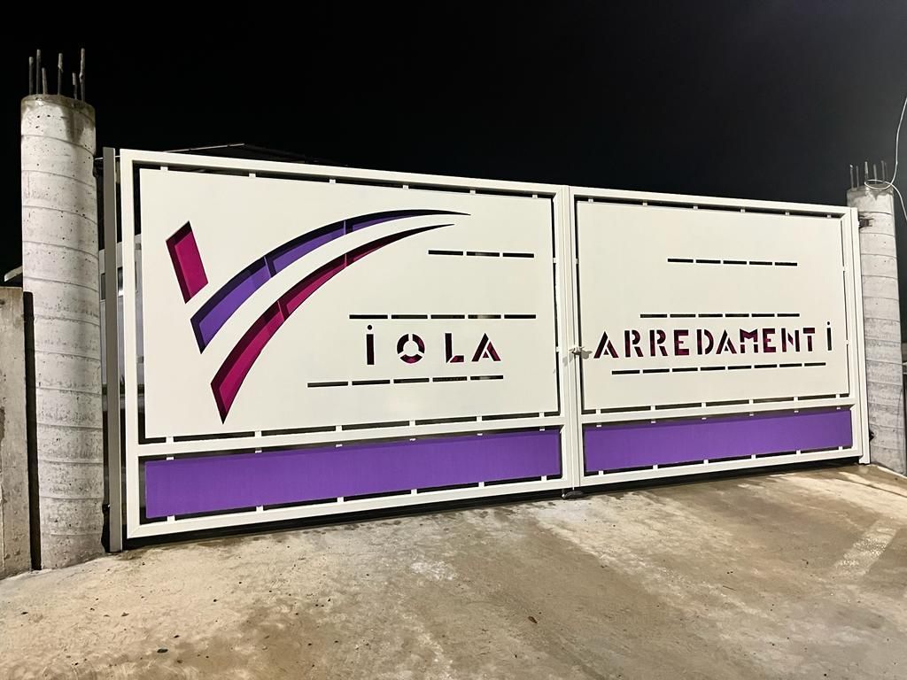 Cancello bianco con logo viola e nero e scritta “TOLA ARREDAMENTI” di notte