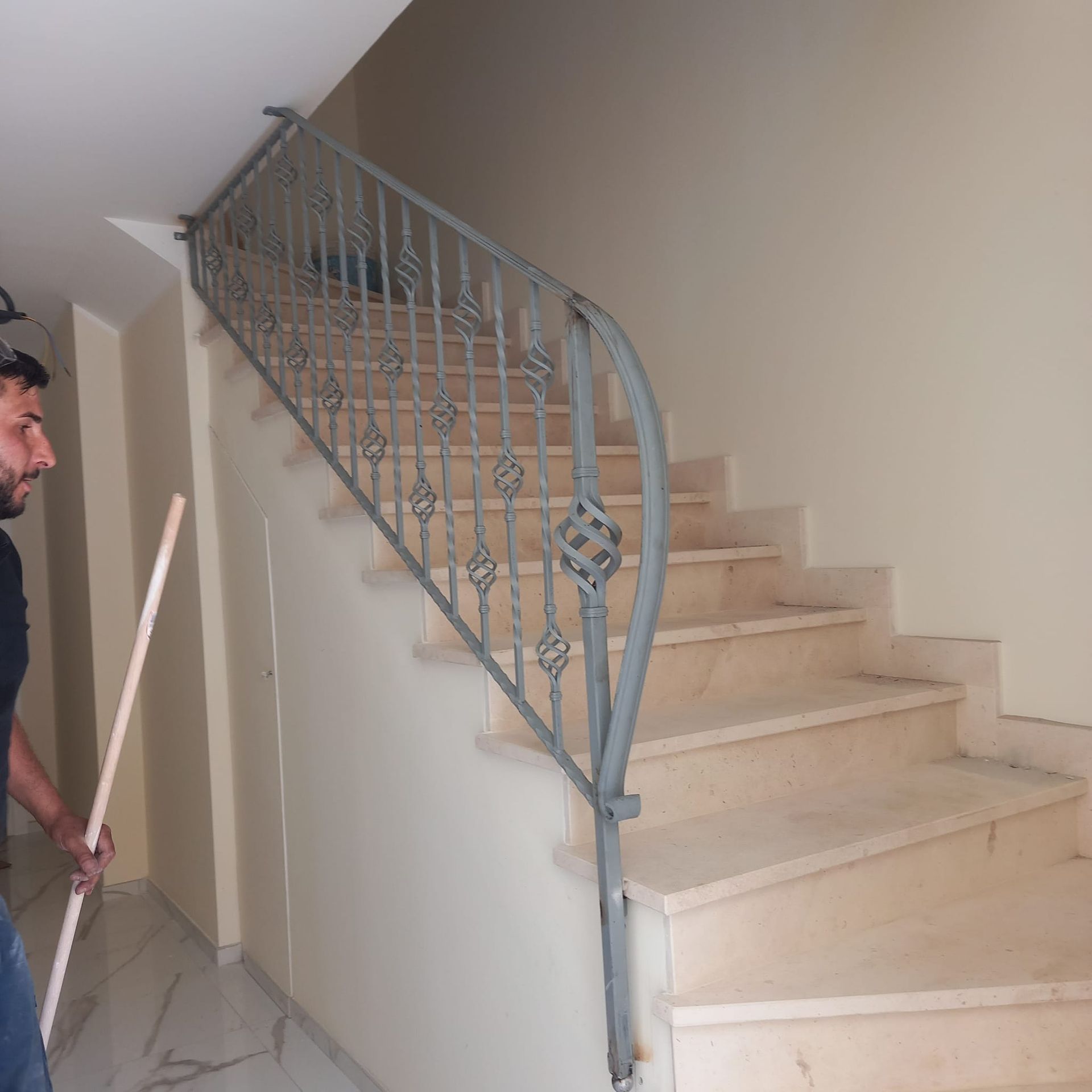Scala interna con gradini beige e ringhiera in metallo grigio decorata; un uomo parzialmente visibile sulla sinistra tiene in mano una scopa.