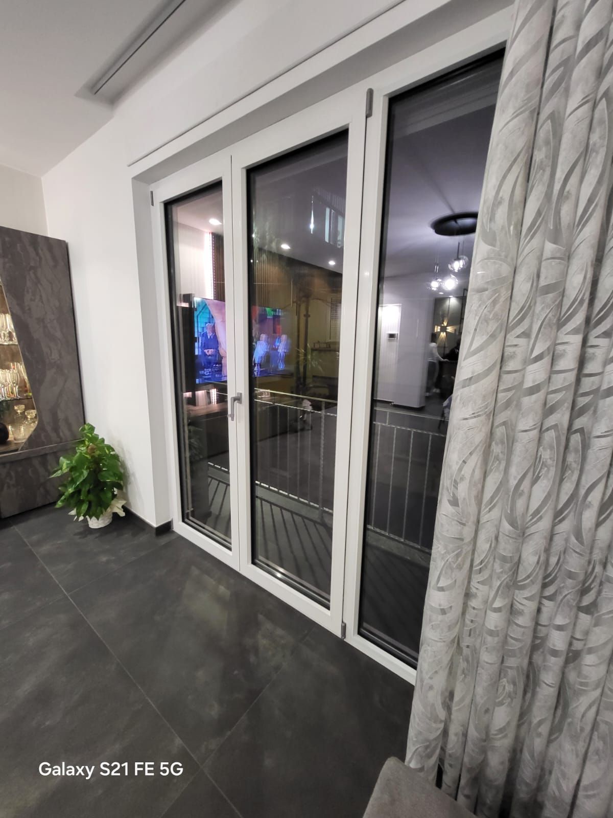 Di notte, delle porte finestre bianche con tende trasparenti si aprono su un balcone, da cui si possono scorgere le luci della città.
