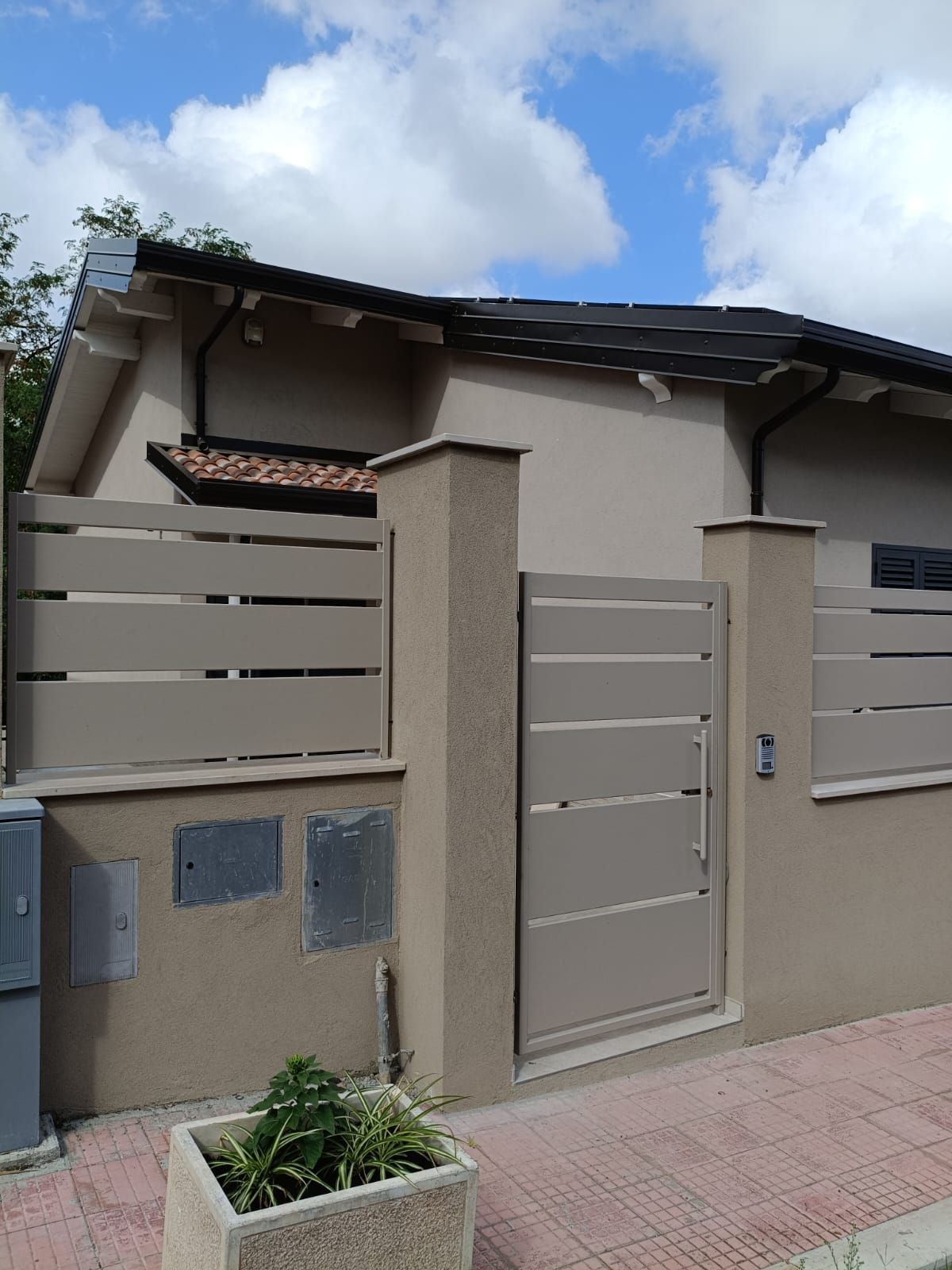 Ingresso di una casa moderna color beige con cancello grigio, muro, piccola fioriera e cielo azzurro sopra.