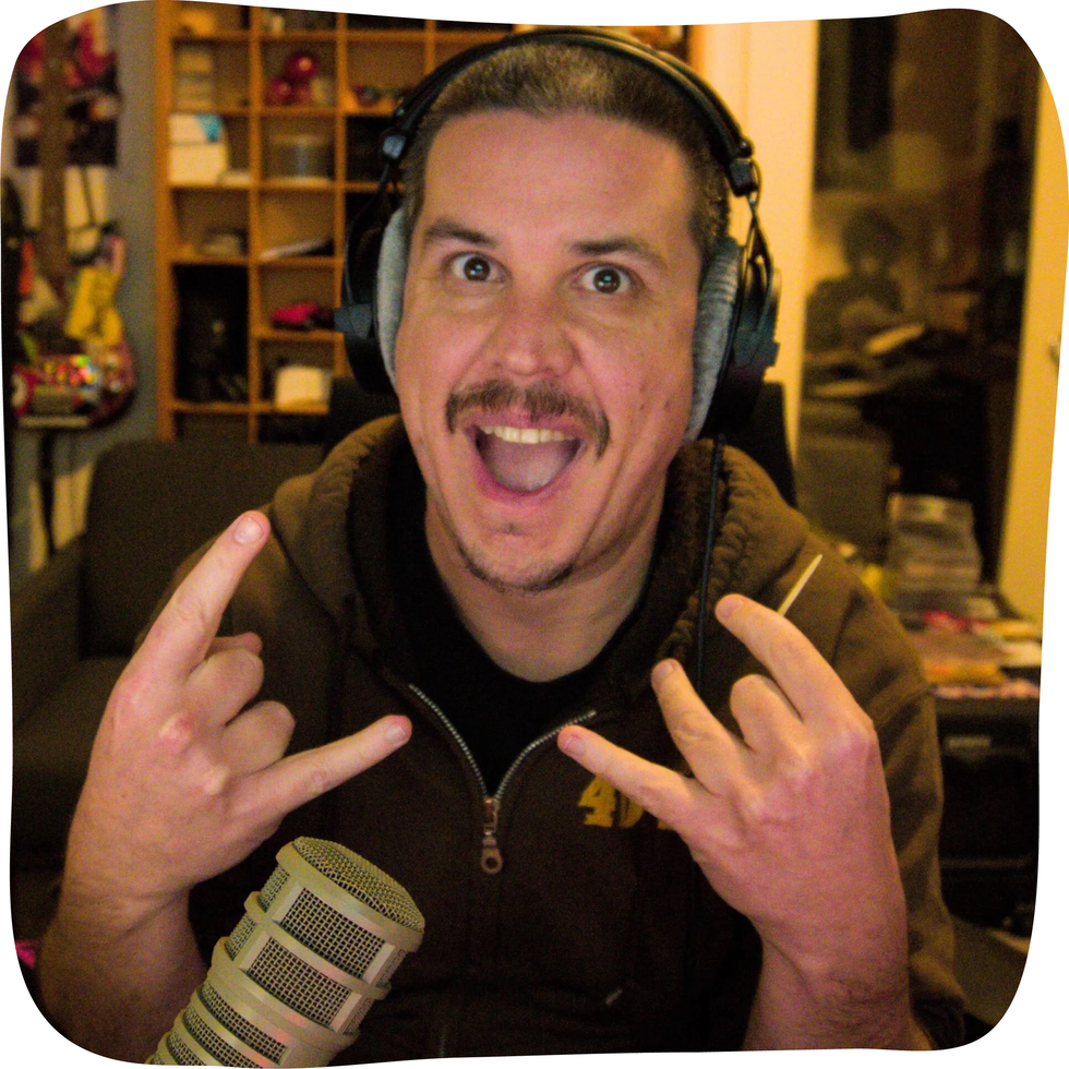 Man with headphones, smiling, giving rock on hand gestures. Sitting in a studio, microphone visible.