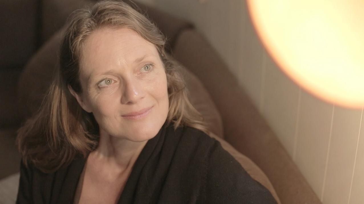 Woman with light brown hair smiles, looking to the right. Soft lighting, indoors.