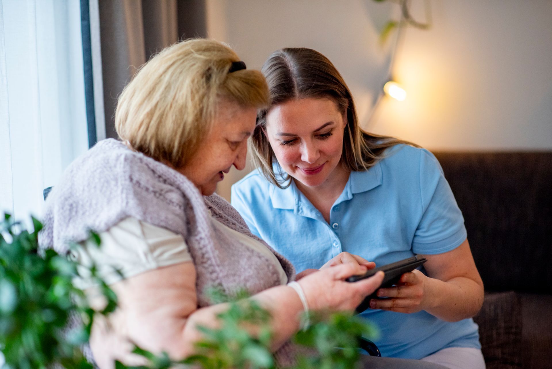 Two women smiling together on a couch, looking at a smartphone in a cozy living room.
