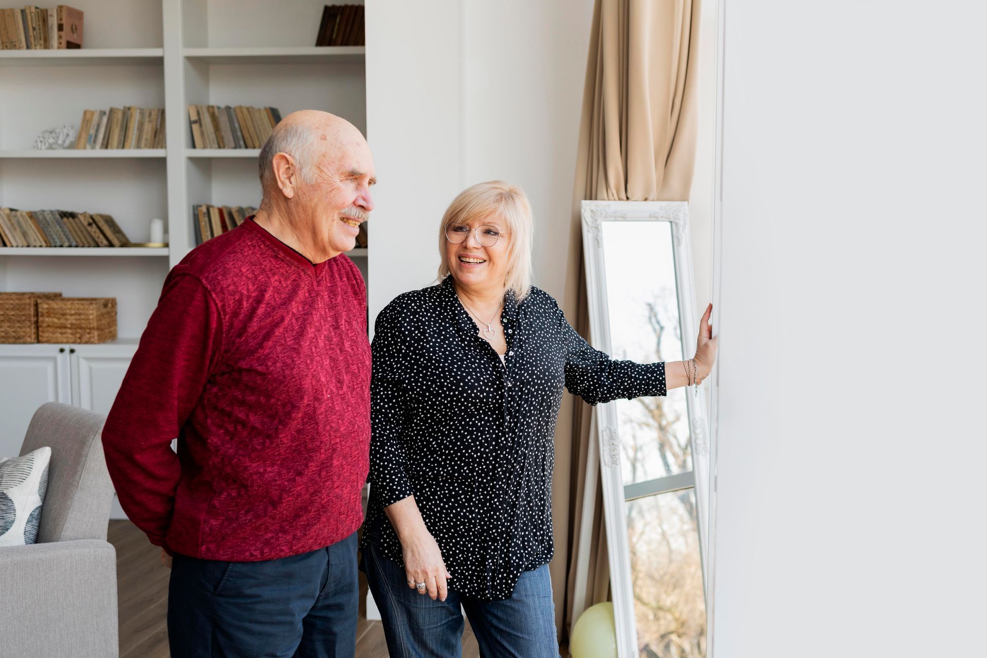 Two people smiling in a bright living room beside a full-length mirror