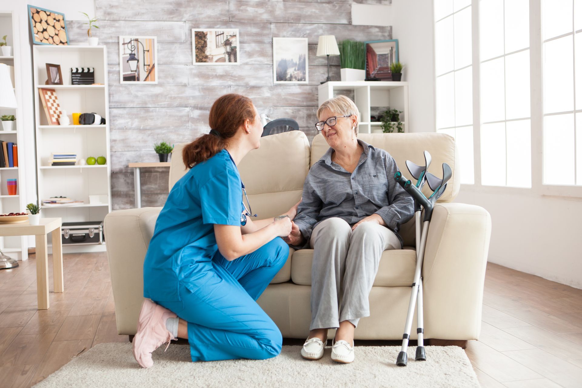 Caregiver in blue scrubs talking with an elderly woman on a sofa in a bright living room with a cane nearby
