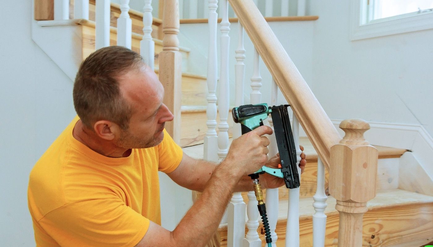 Man installing trim on a staircase with a nail gun, wearing a yellow shirt.