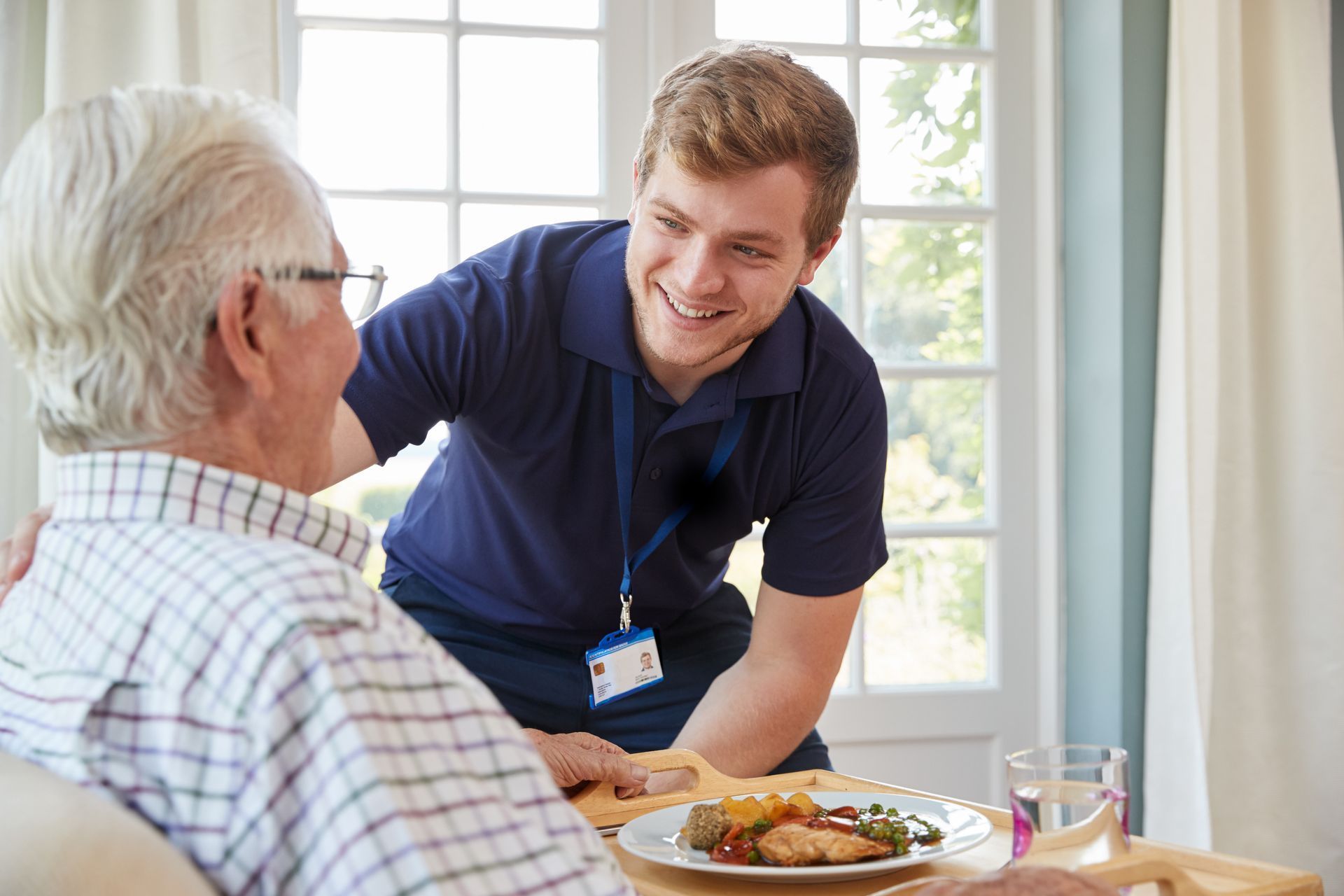 Caregiver assists senior at a dining table. Both are smiling, near a window with food and drink.