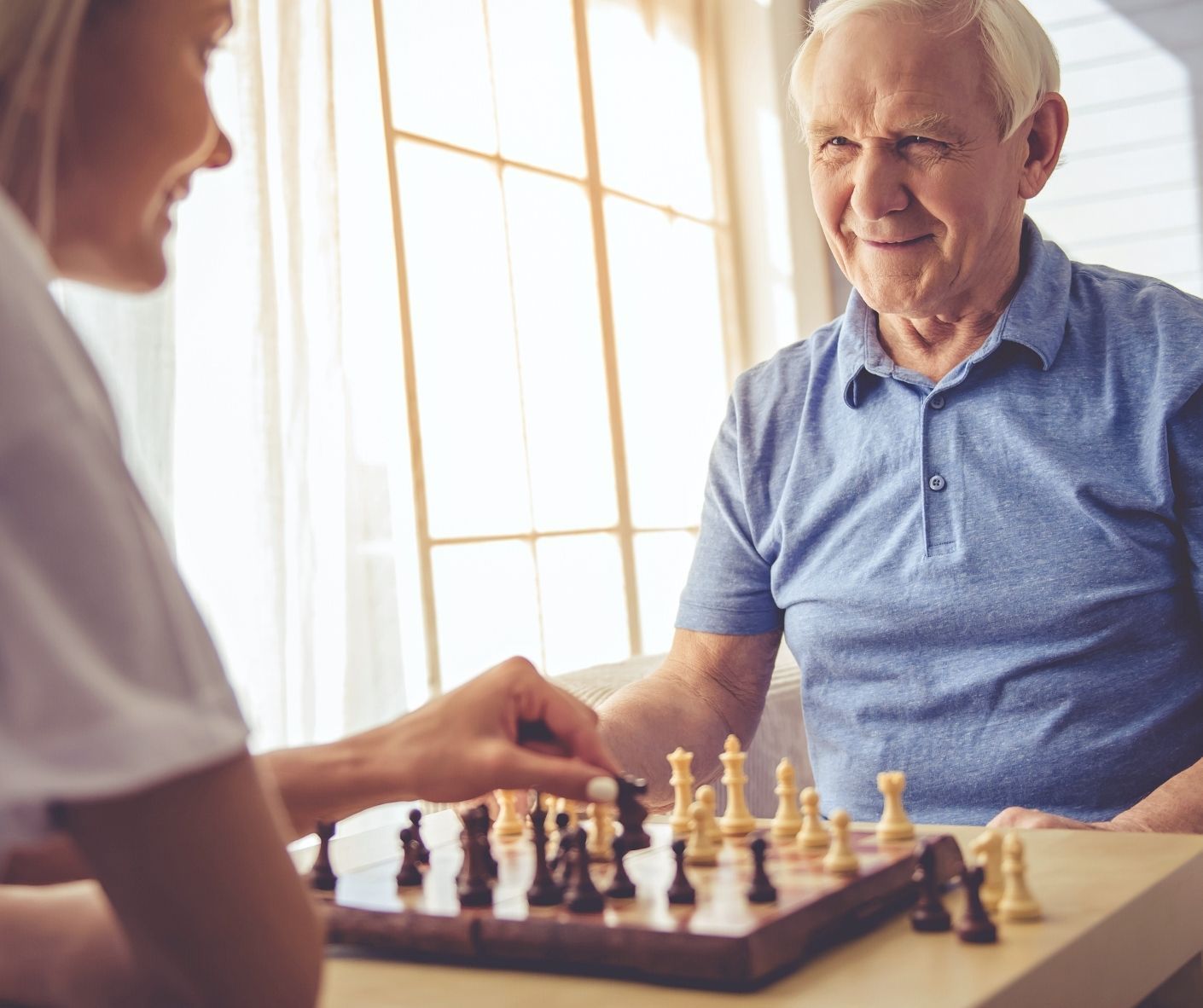 Two people playing chess at a sunlit table, with an elderly man smiling across the board.