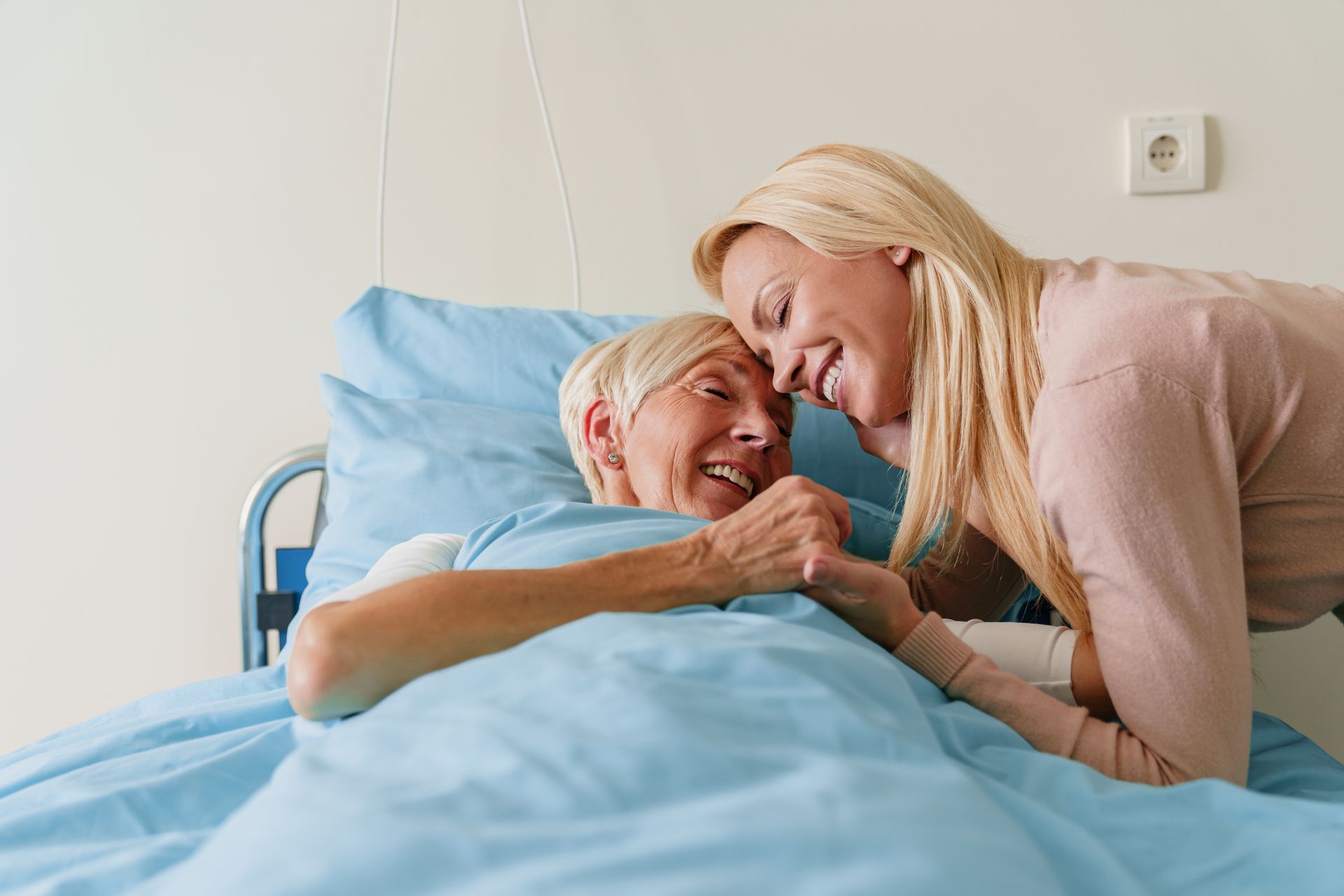 Woman comforting smiling patient in hospital bed, holding hands under a blue blanket