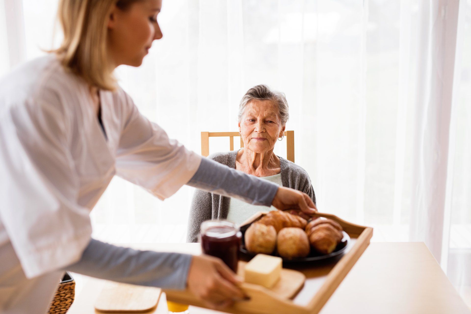 Caregiver serving breakfast to a seated older adult at a table. Croissants, jelly, butter on tray.