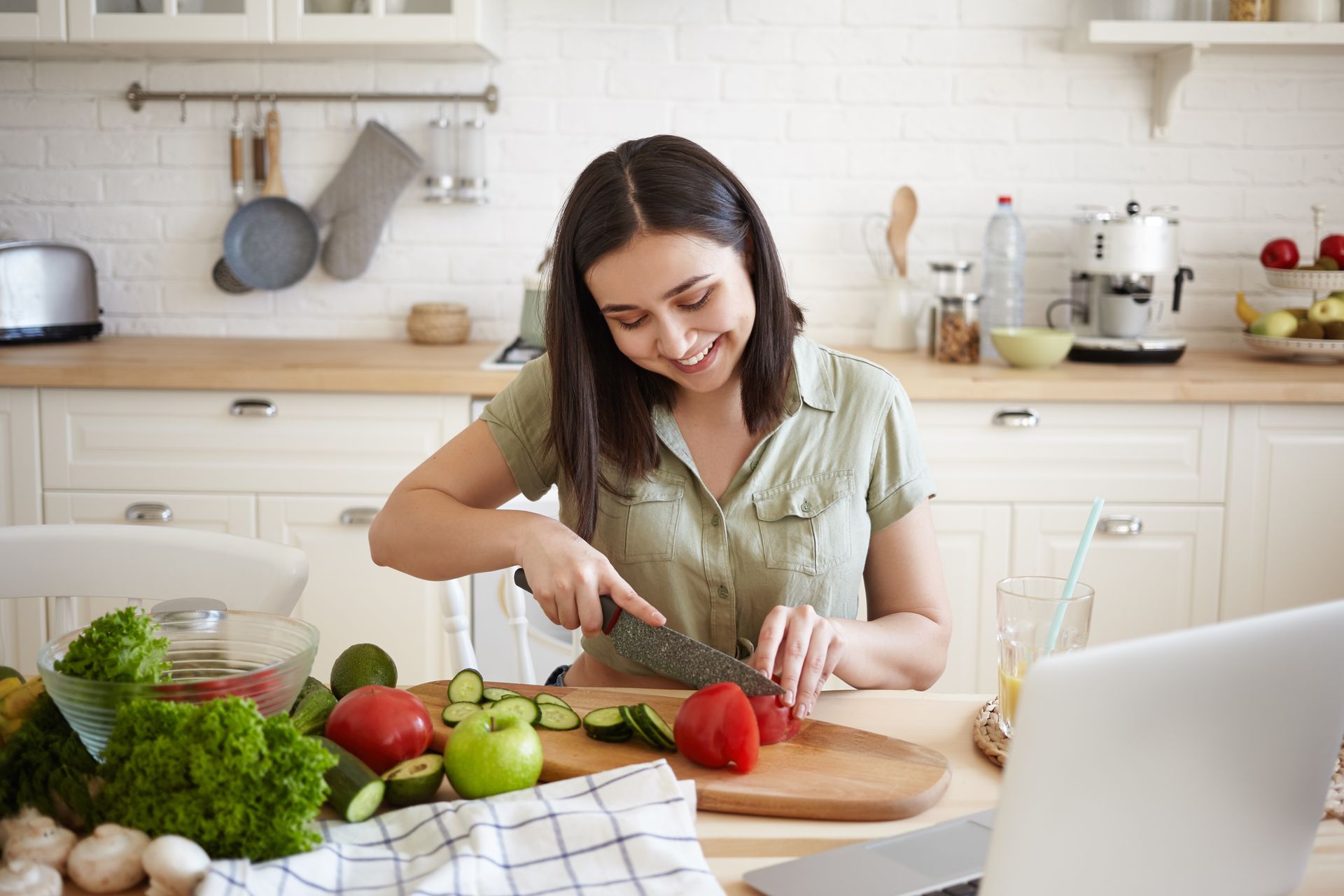 A woman is cutting vegetables in a kitchen in front of a laptop.