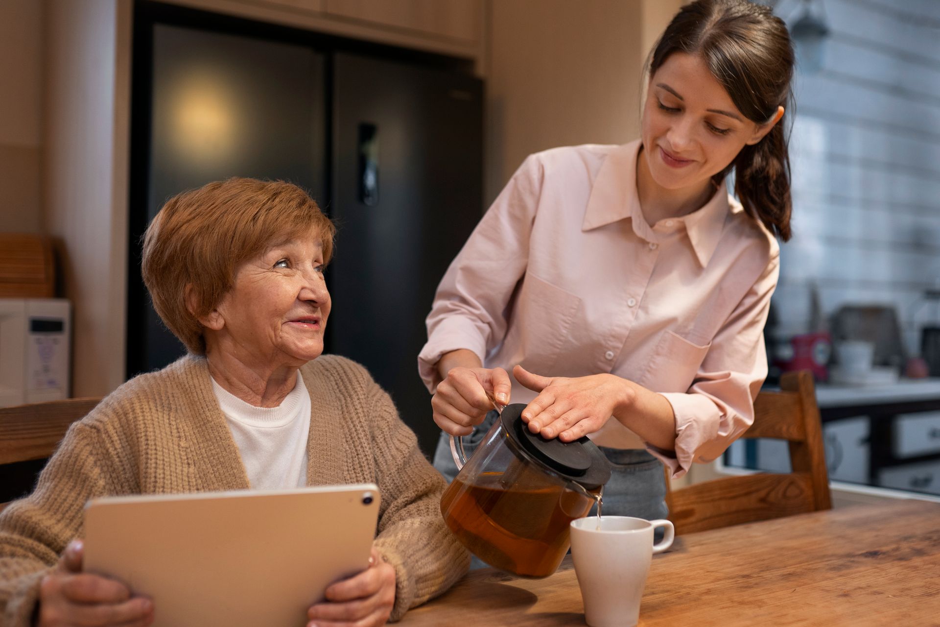 A woman is pouring tea into a cup while an elderly woman looks at a tablet.