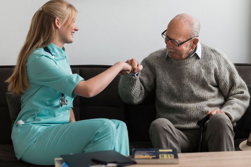 Caregiver in teal scrubs gently fist-bumps an elderly man seated in a living room