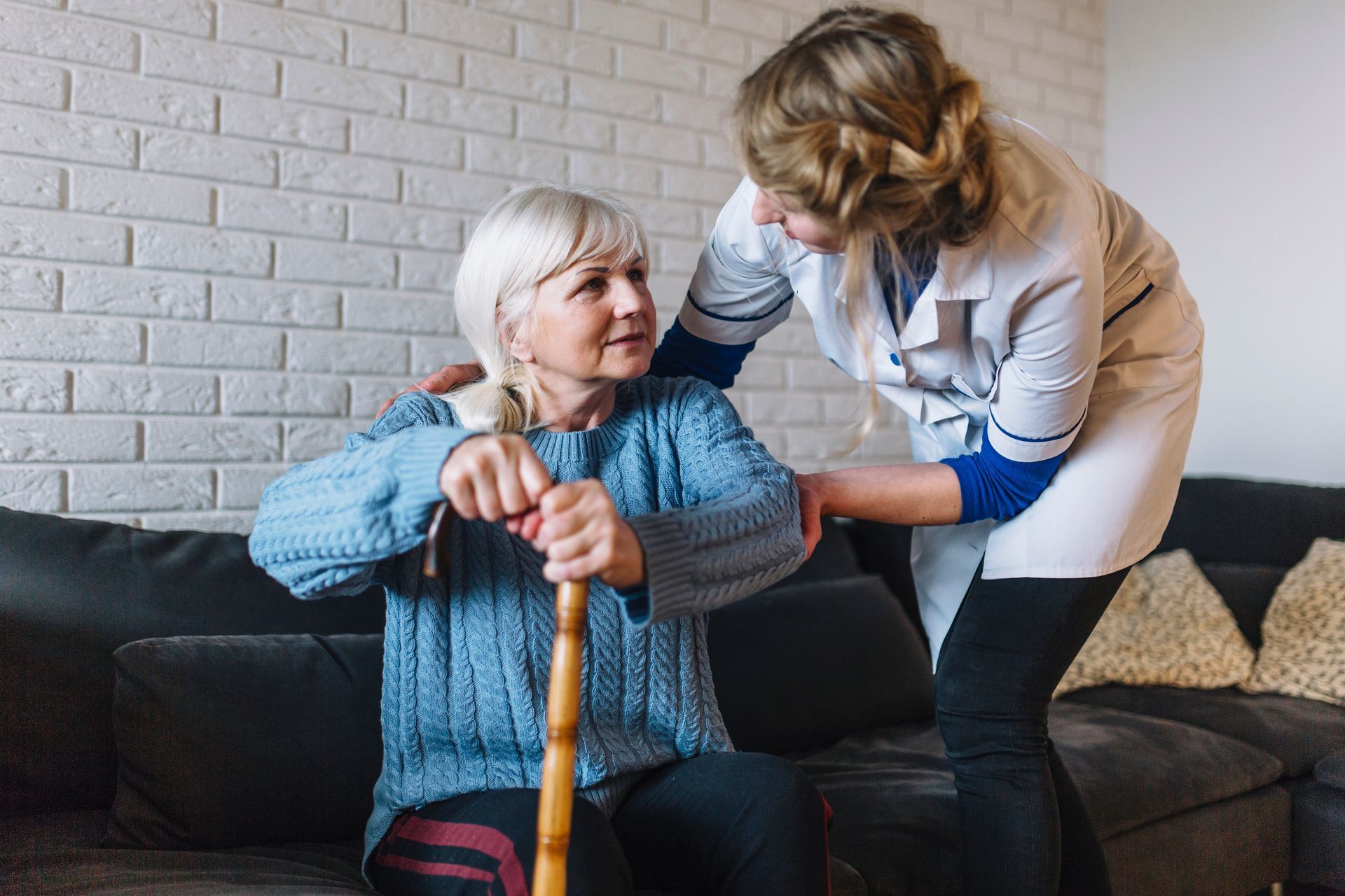 An elderly woman is sitting on a couch with a cane and a nurse is helping her.