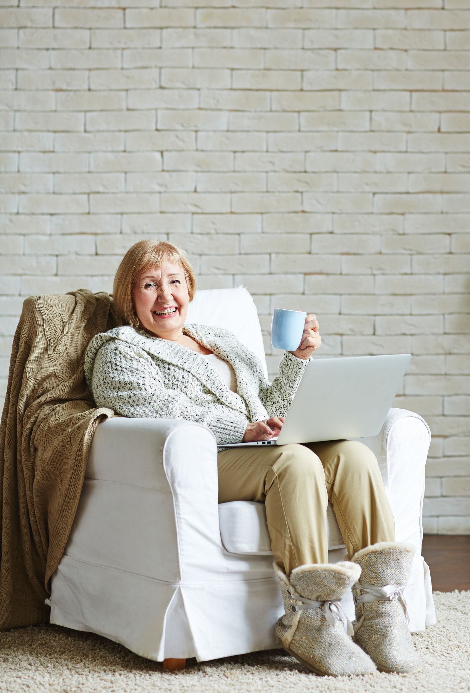 An elderly woman is sitting in a chair with a laptop and a cup of coffee.