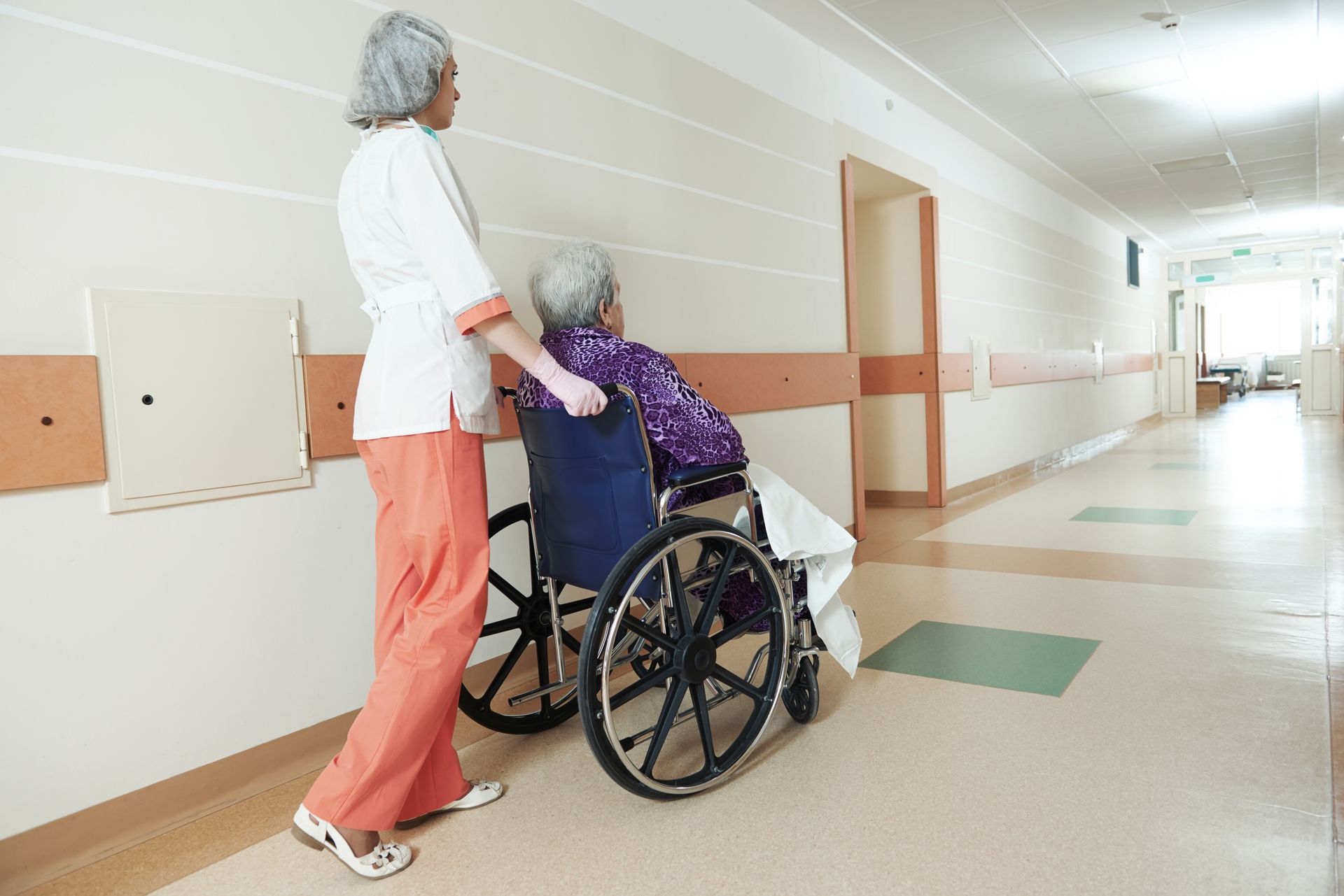 A nurse pushes an elderly woman in a wheelchair down a hospital hallway.
