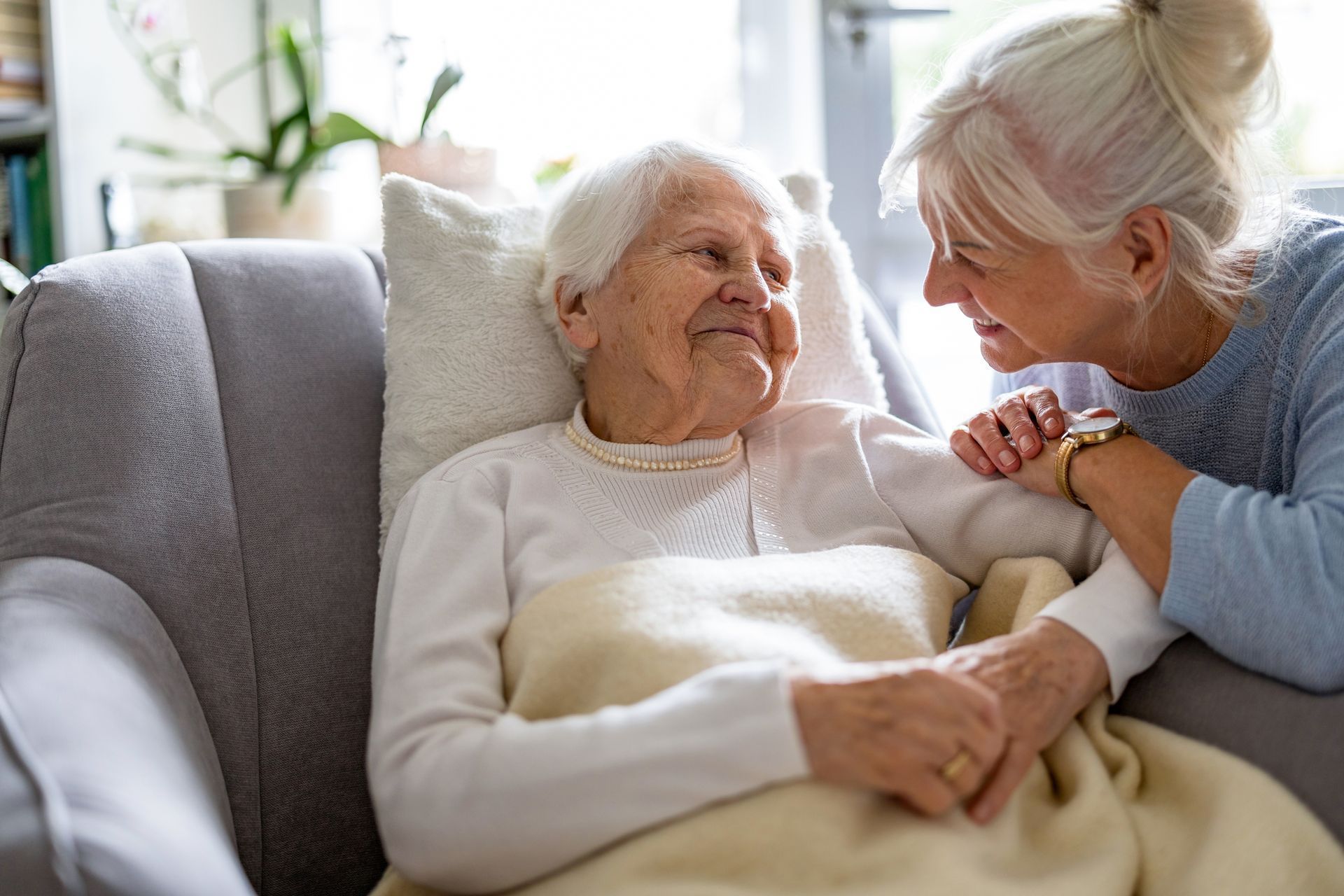 Older woman in a chair is comforted by another woman who holds her hand indoors.