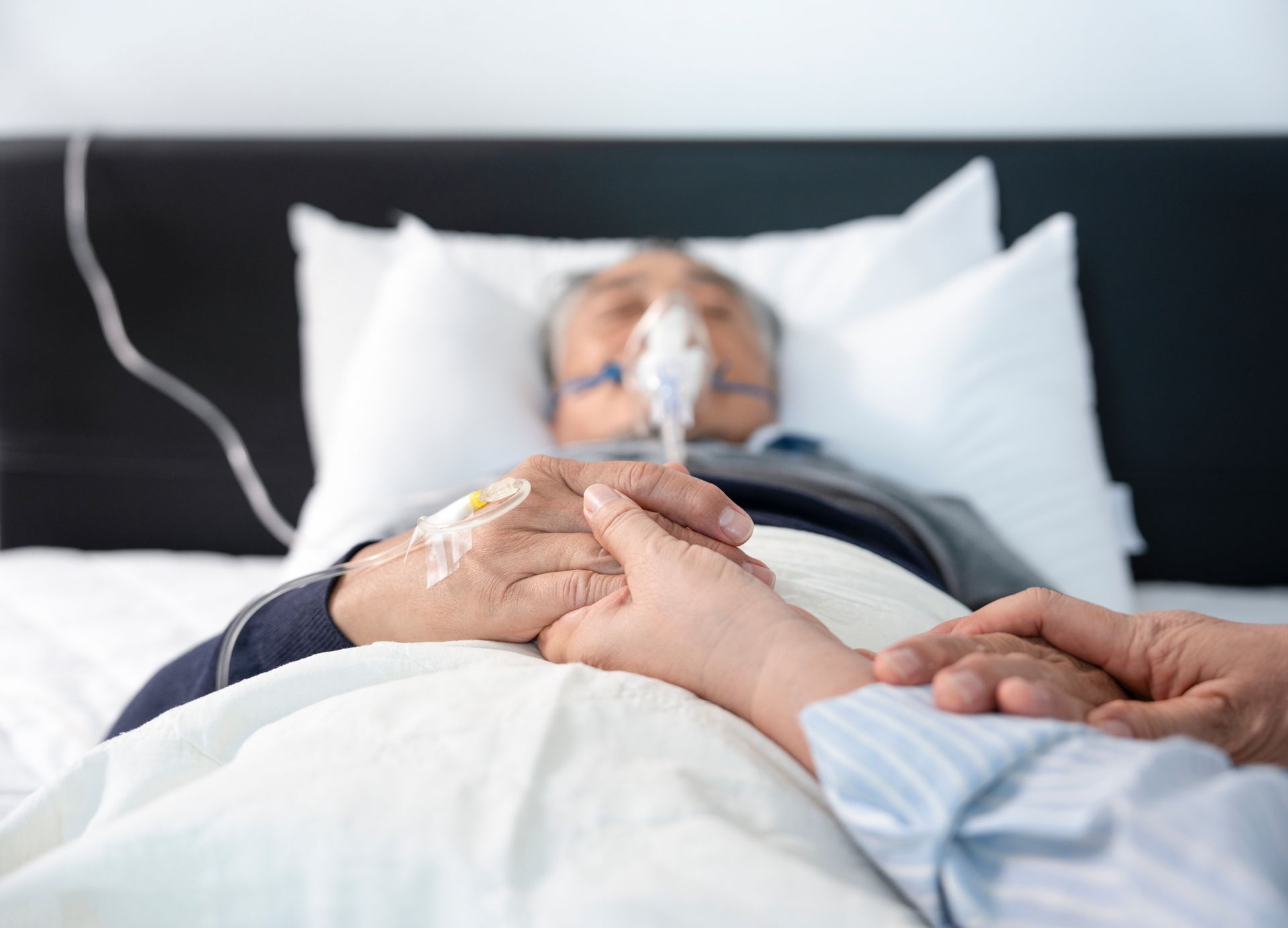 Person in bed with oxygen mask, hand held by another person.