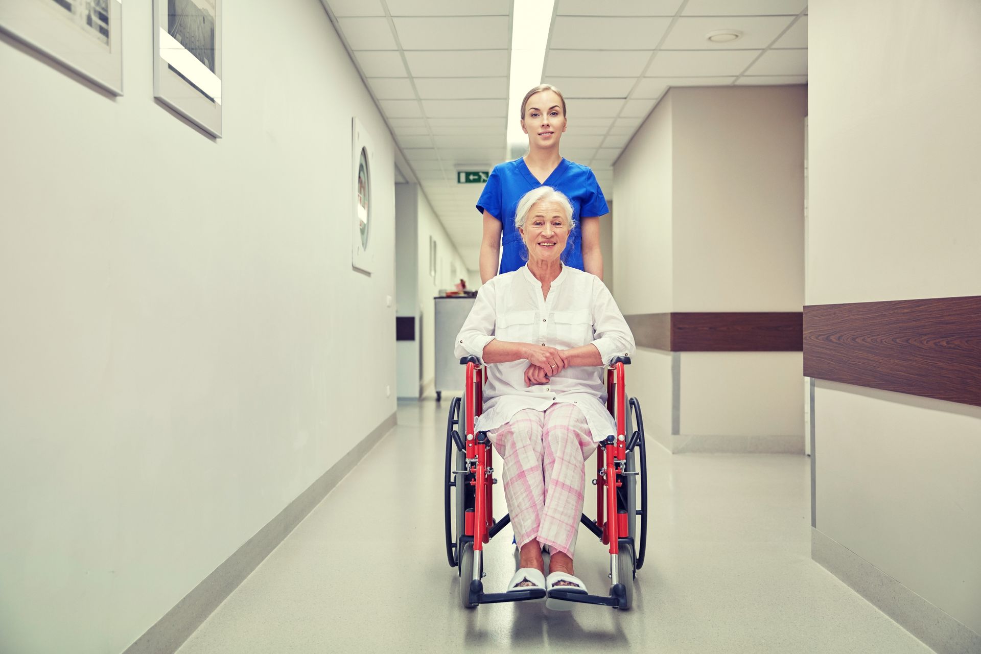 A nurse pushes an elderly woman in a wheelchair down a hospital hallway.