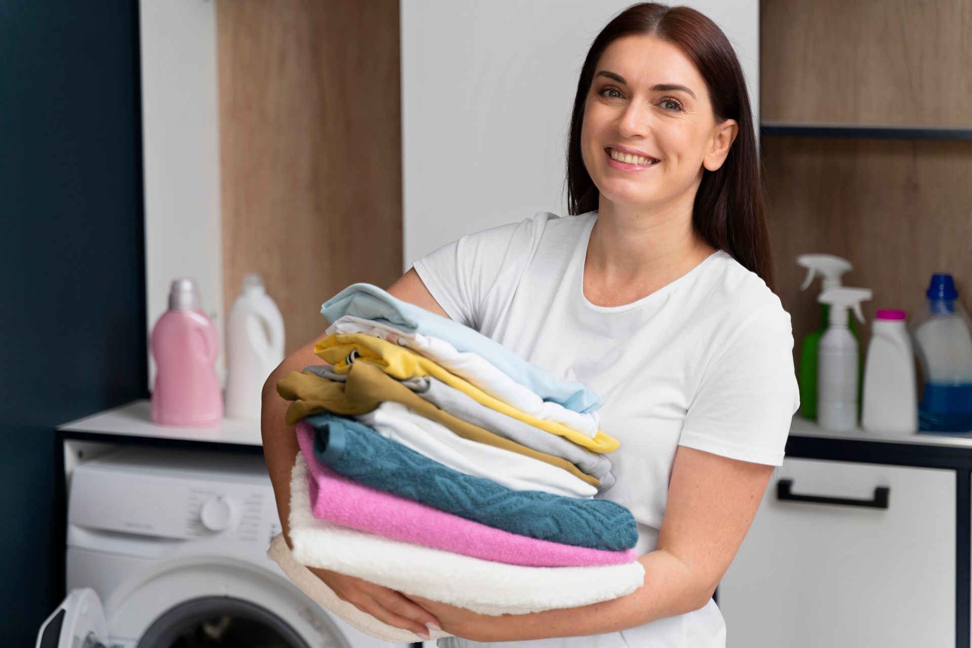 A woman is holding a pile of clothes in front of a washing machine.