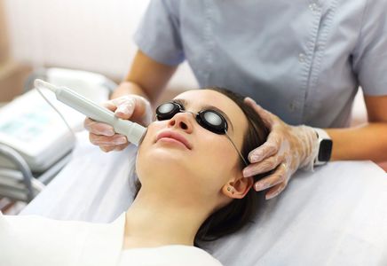 A professional provides a facial treatment to a client wearing protective goggles while lying on a clinic bed.