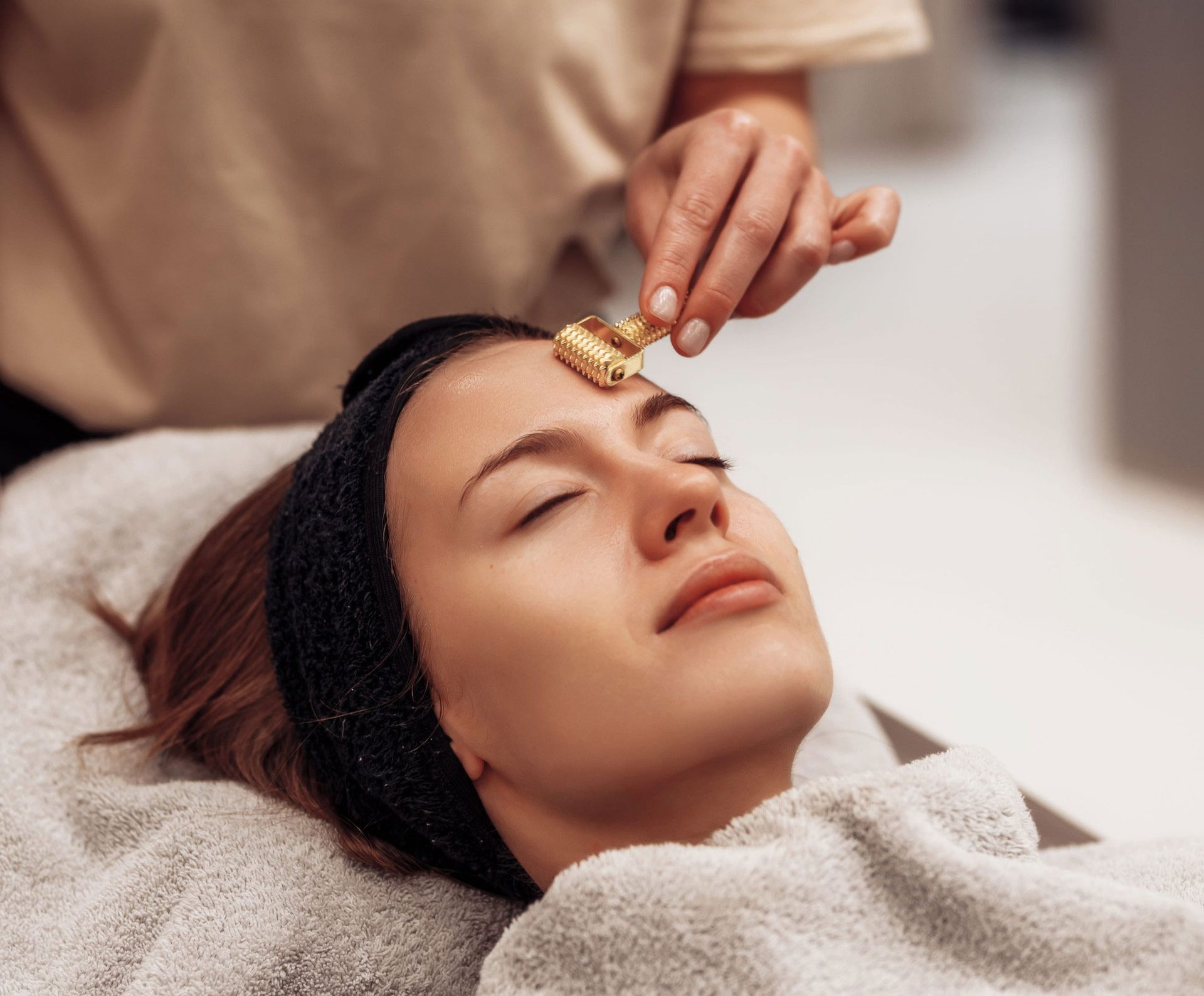 A woman with eyes closed lies on a white surface next to a palm leaf, purple bath salts, and lit white candles.