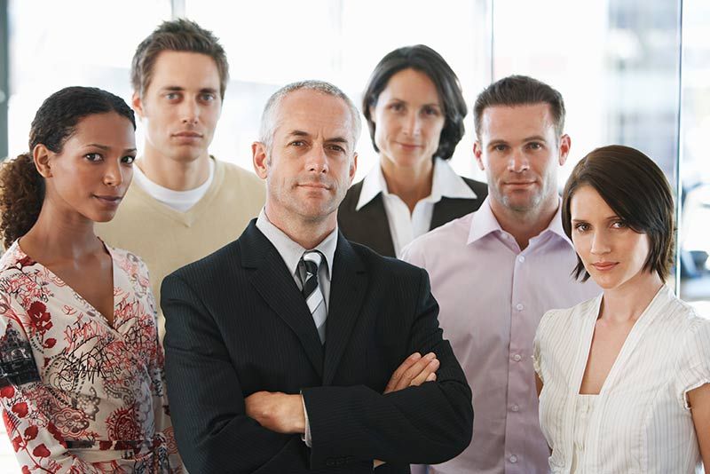 A group of professionals in business attire stand together in an office setting, looking toward the camera.