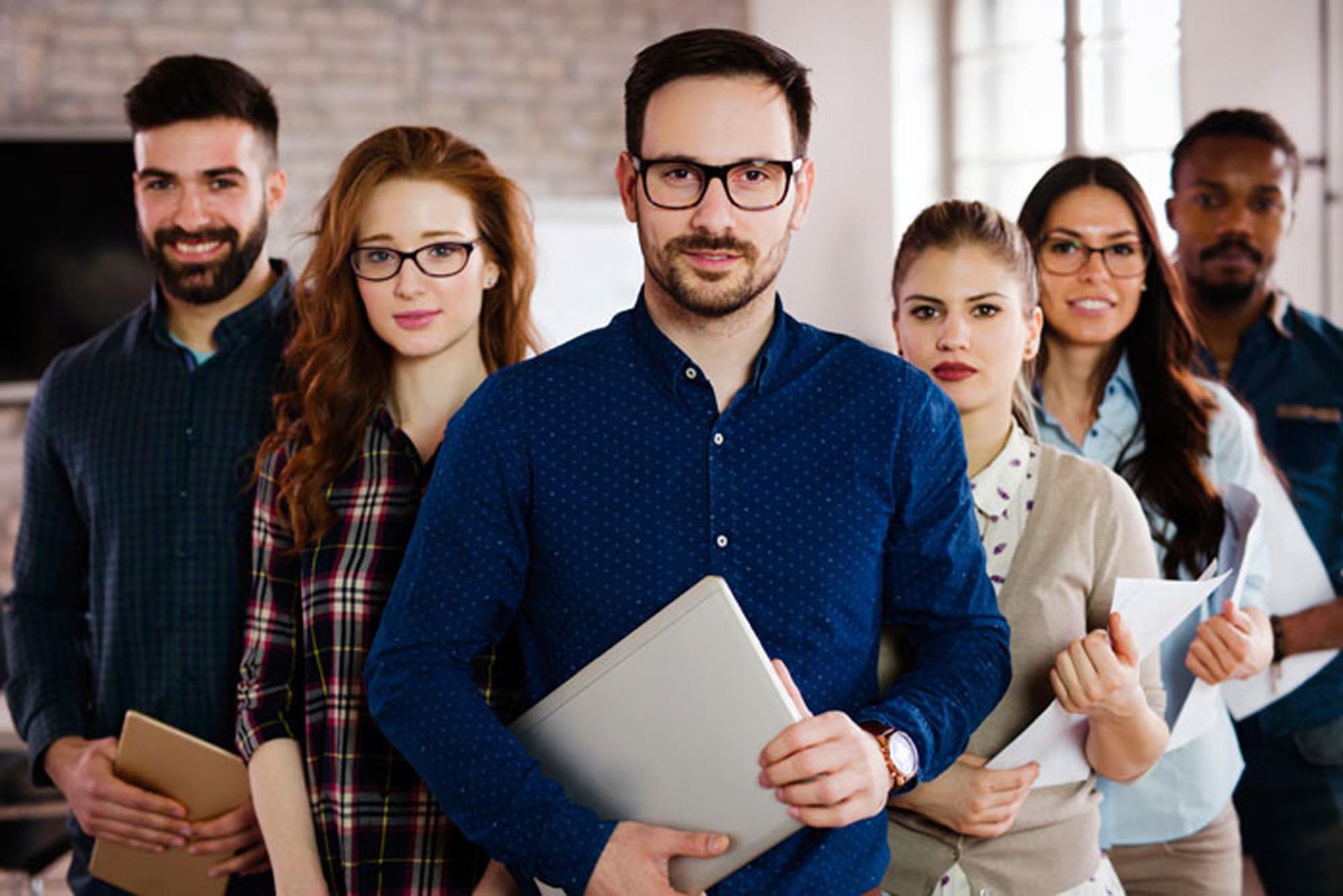 A diverse group of six professionals standing in a line, holding notebooks and documents, looking at the camera.