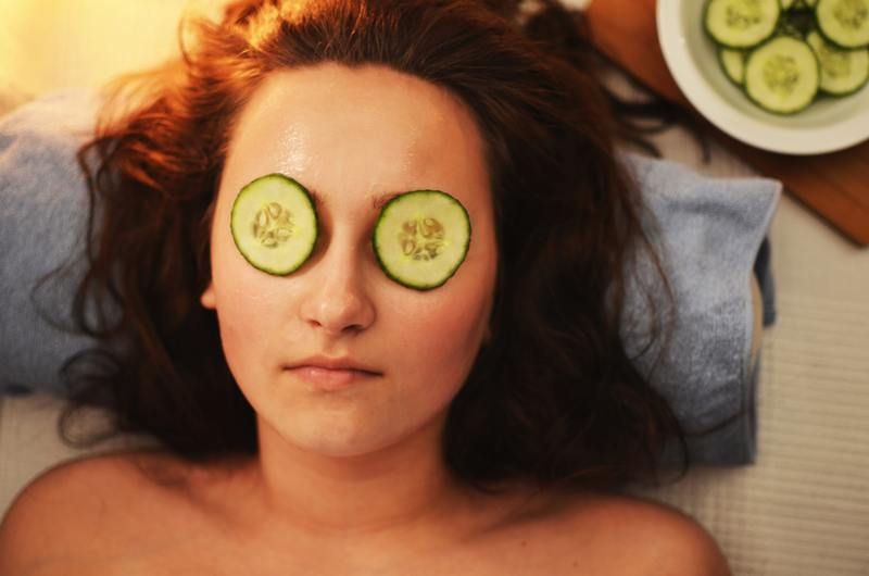 A person lying down with cucumber slices on their eyes, with a bowl of cucumber slices nearby.