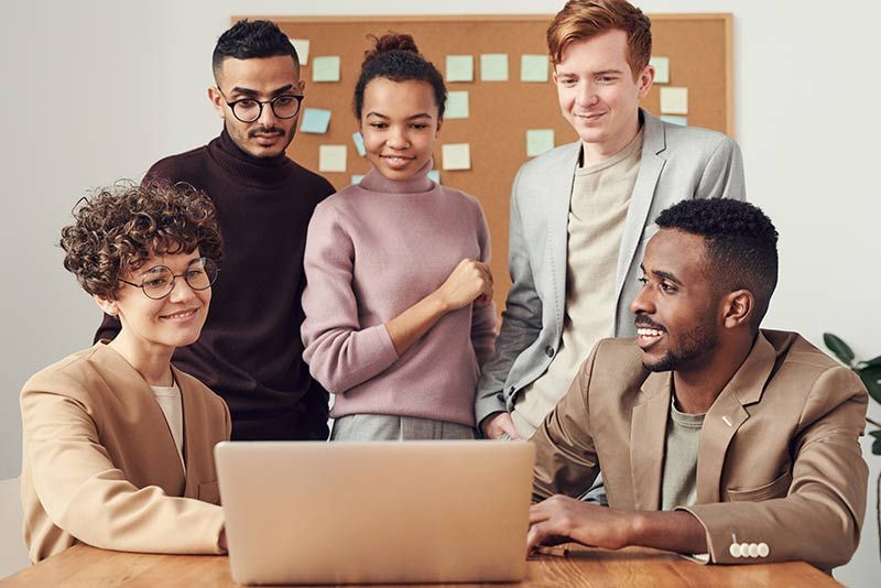 A diverse group of colleagues gather around a laptop in an office, looking at the screen and smiling during a discussion.
