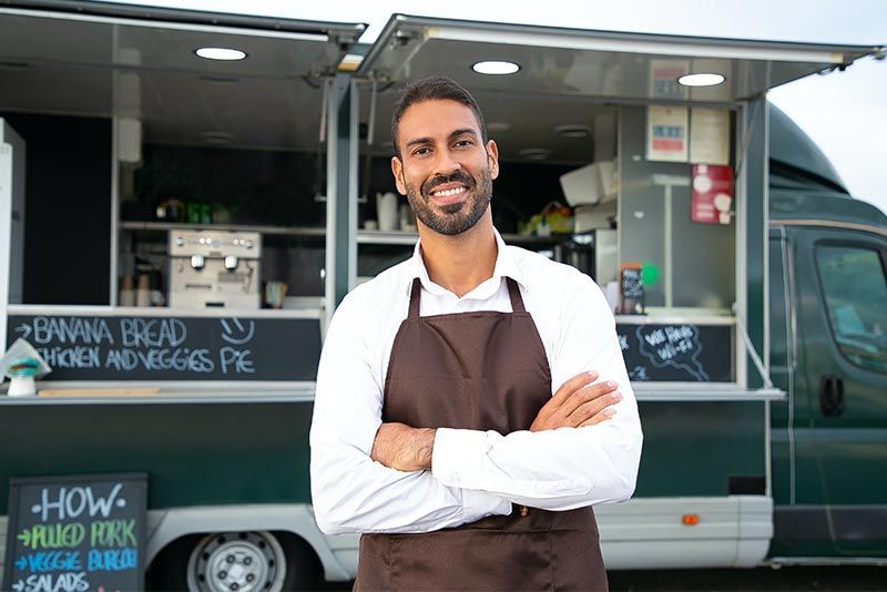 A smiling person in a white shirt and brown apron with arms crossed stands in front of a food truck with chalkboard menus.