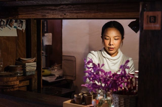 A person in a white turtleneck works behind a wooden counter filled with small cups and a vase of purple orchids.