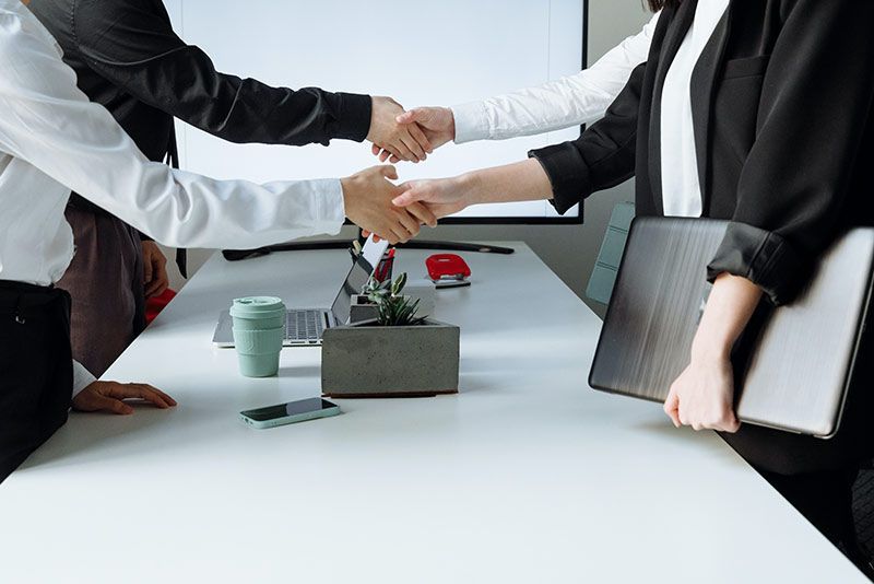 Two people in business attire shake hands over a conference table in an office setting.