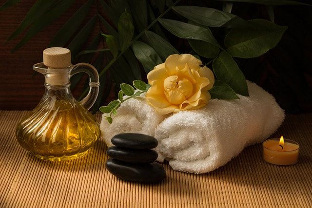 Spa setting with massage oil in a glass carafe, rolled white towels, a yellow flower, stacked black stones, and a candle.