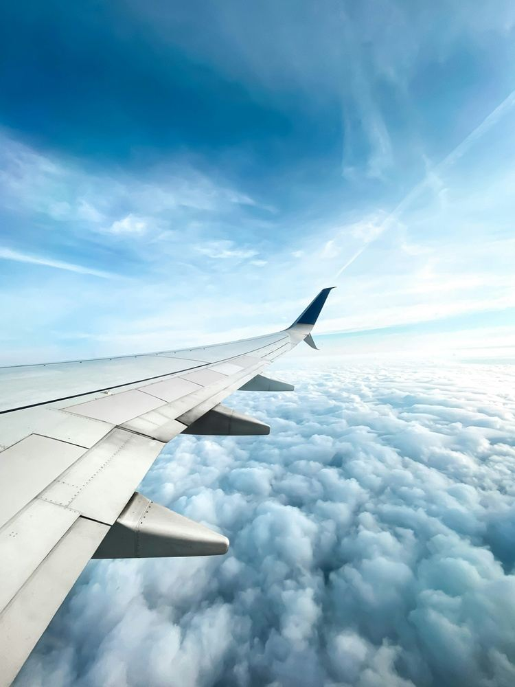 Airplane wing soaring above a fluffy cloud layer in a blue sky.