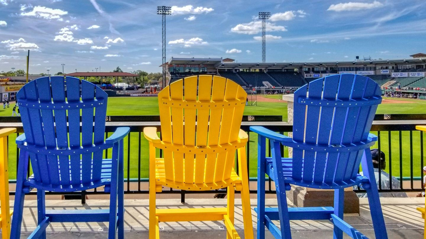 Three colorful Adirondack chairs overlook a baseball field on a sunny day.
