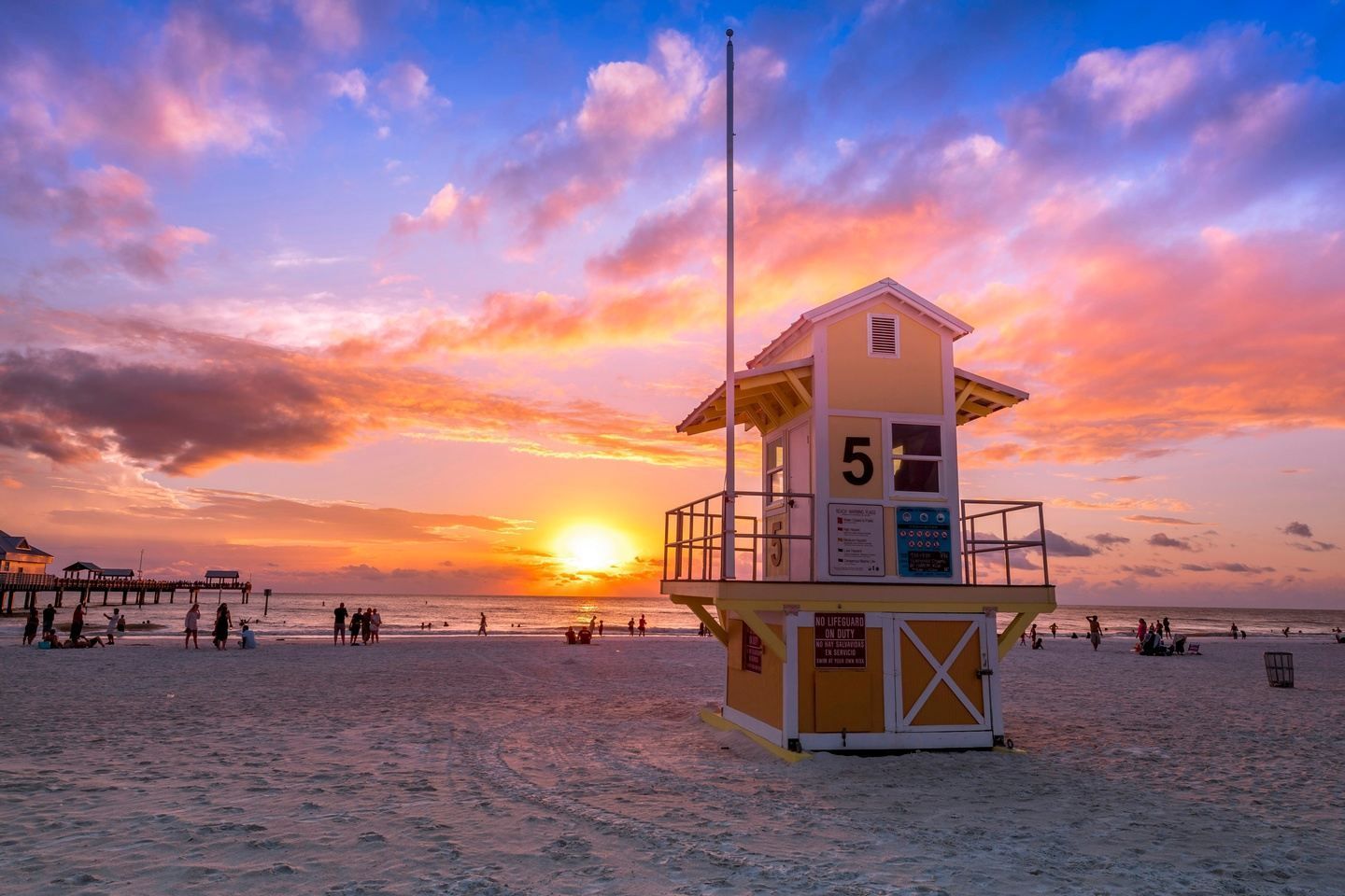 Sunset at the beach with a yellow lifeguard stand, silhouetted people, and colorful clouds.