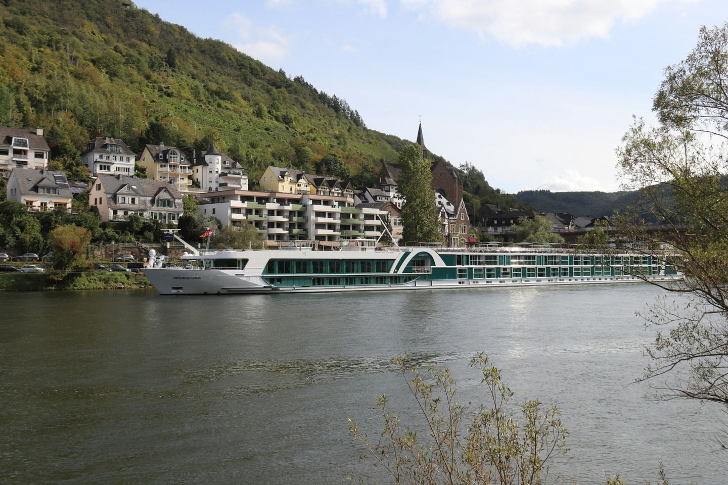 River cruise ship sailing past a town on a hillside with green trees; overcast sky.