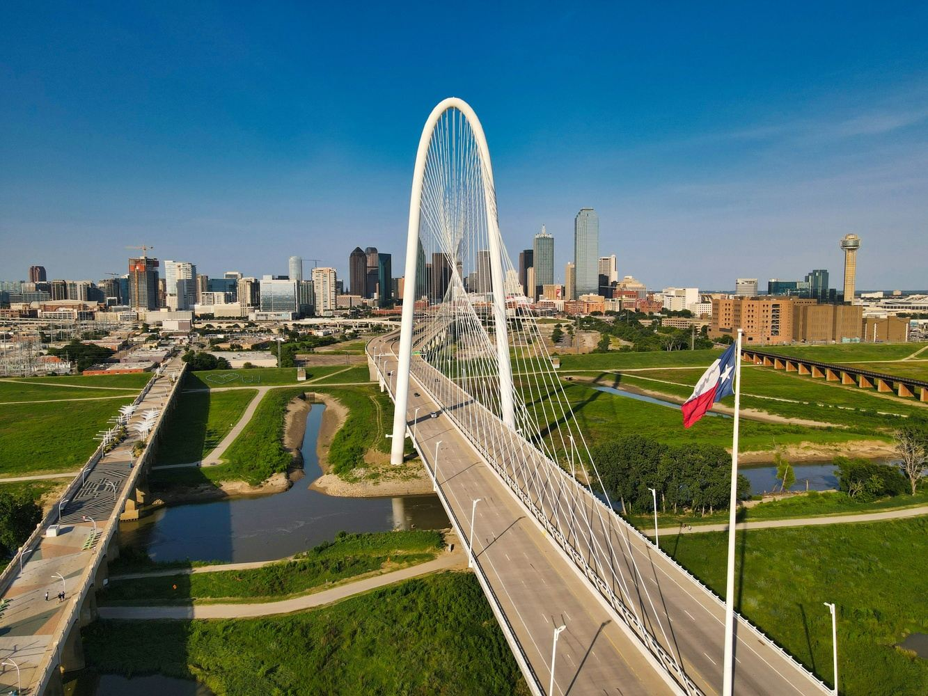 White arch bridge over a river, with Dallas skyline in the background, blue sky, and Texas flag.