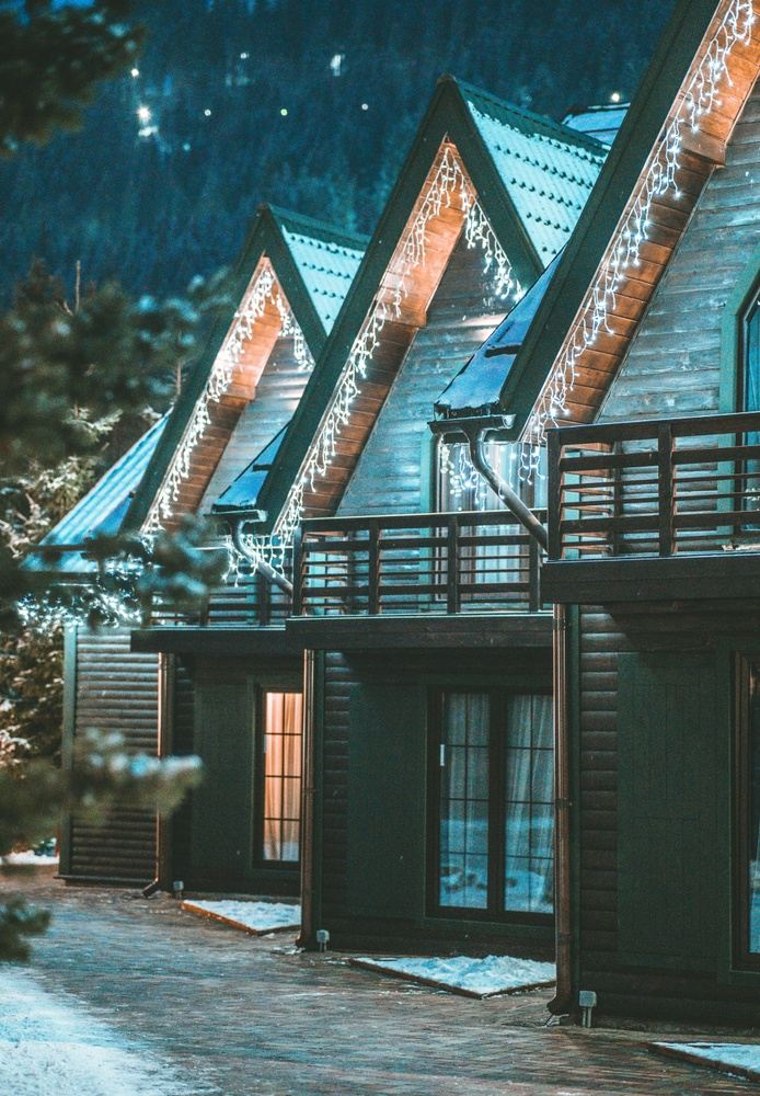 Row of wooden cabins with triangular roofs and Christmas lights, set against a dark, snowy forest.