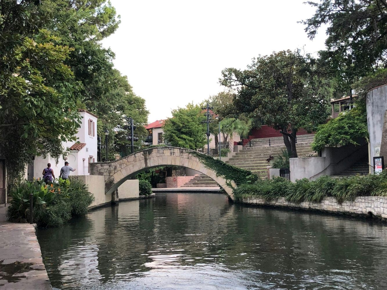 Stone bridge over a calm river in a city park with lush greenery and buildings along the banks.