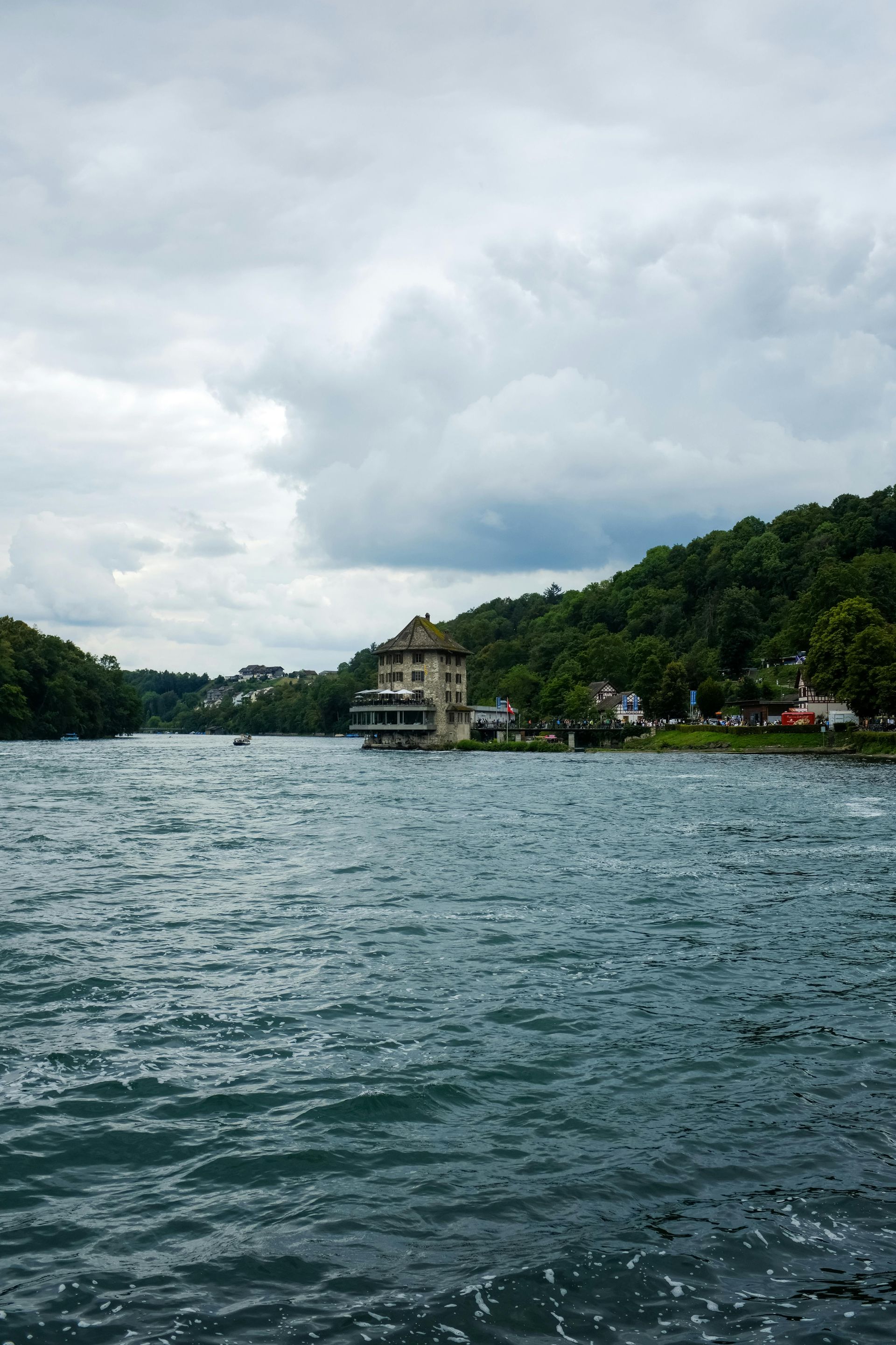 River with rippling water, building on shore, lush green hill under cloudy sky.