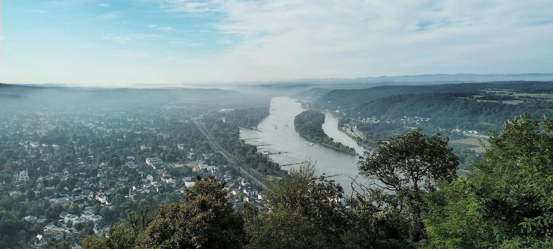Overlook of a river winding through a valley, trees in foreground. Hazy sky, houses and buildings.