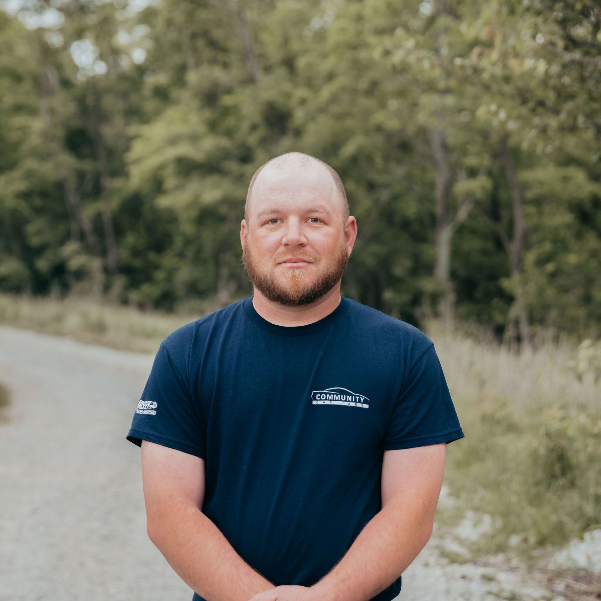 Man standing on a road, wearing a navy T-shirt, with trees and greenery in the background.