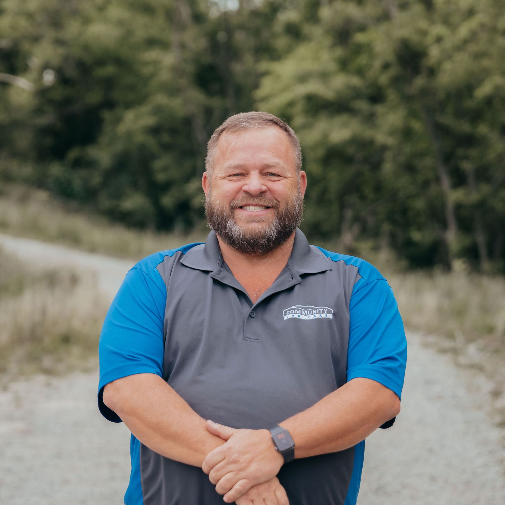 Smiling man in gray and blue shirt standing on a gravel road with trees behind him