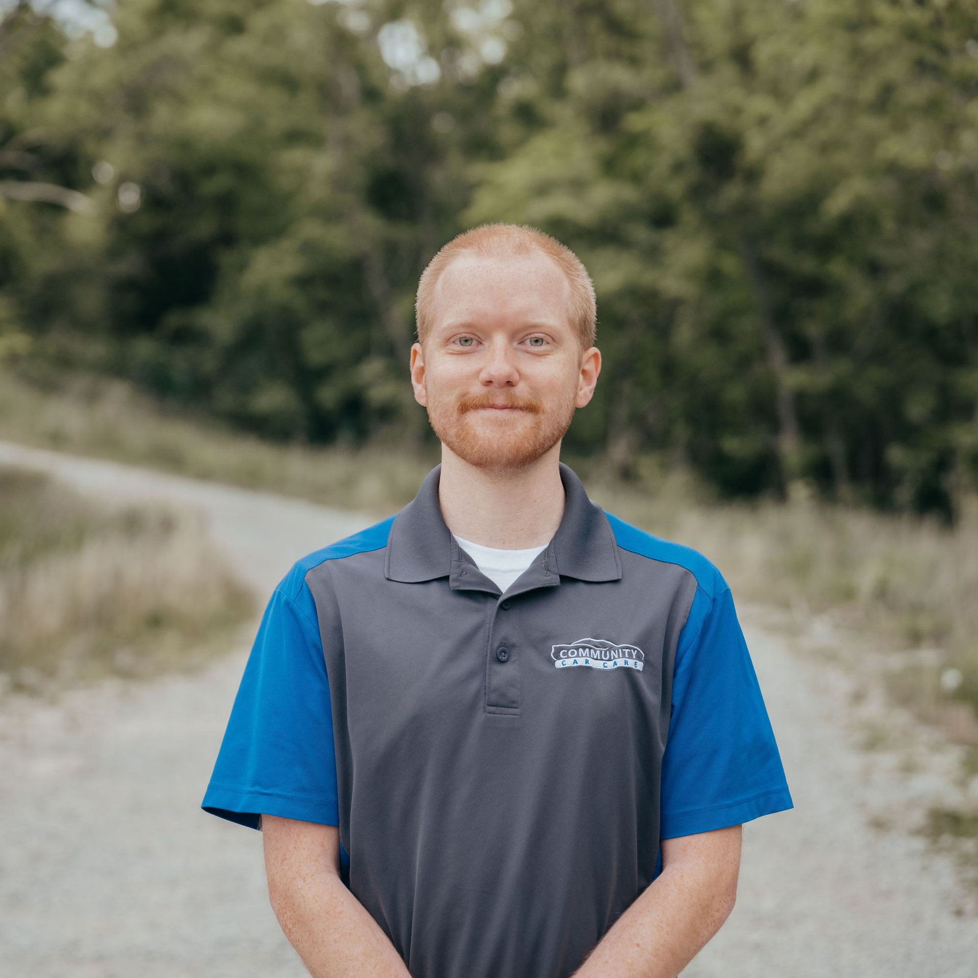 Man in a gray and blue polo shirt standing outdoors with trees blurred in the background